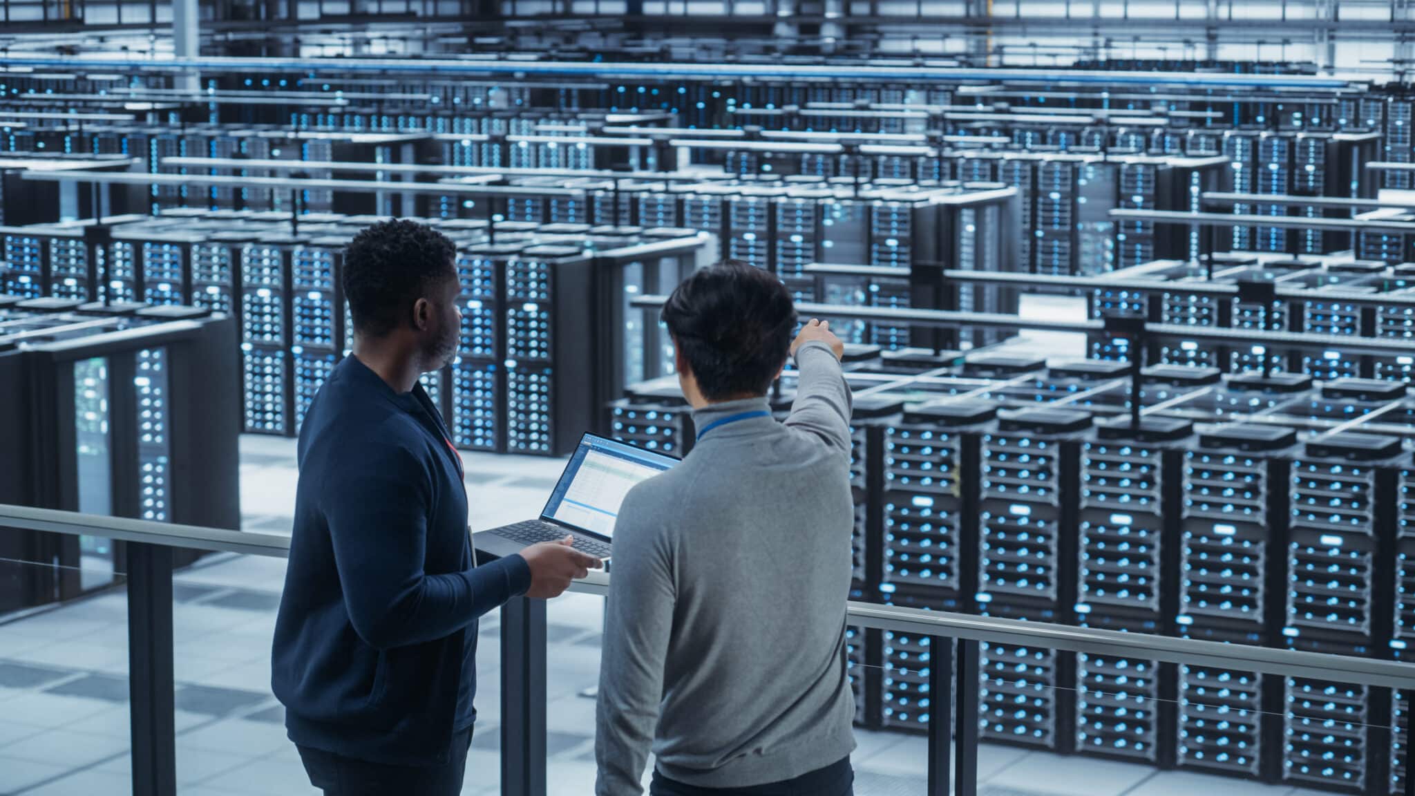 Two Multiethnic Male Data Center System Administrators and IT Specialists Talk, Use Laptop. Information Technology Engineers work on Cyber Security Network Protection in Cloud Computing Server Farm.